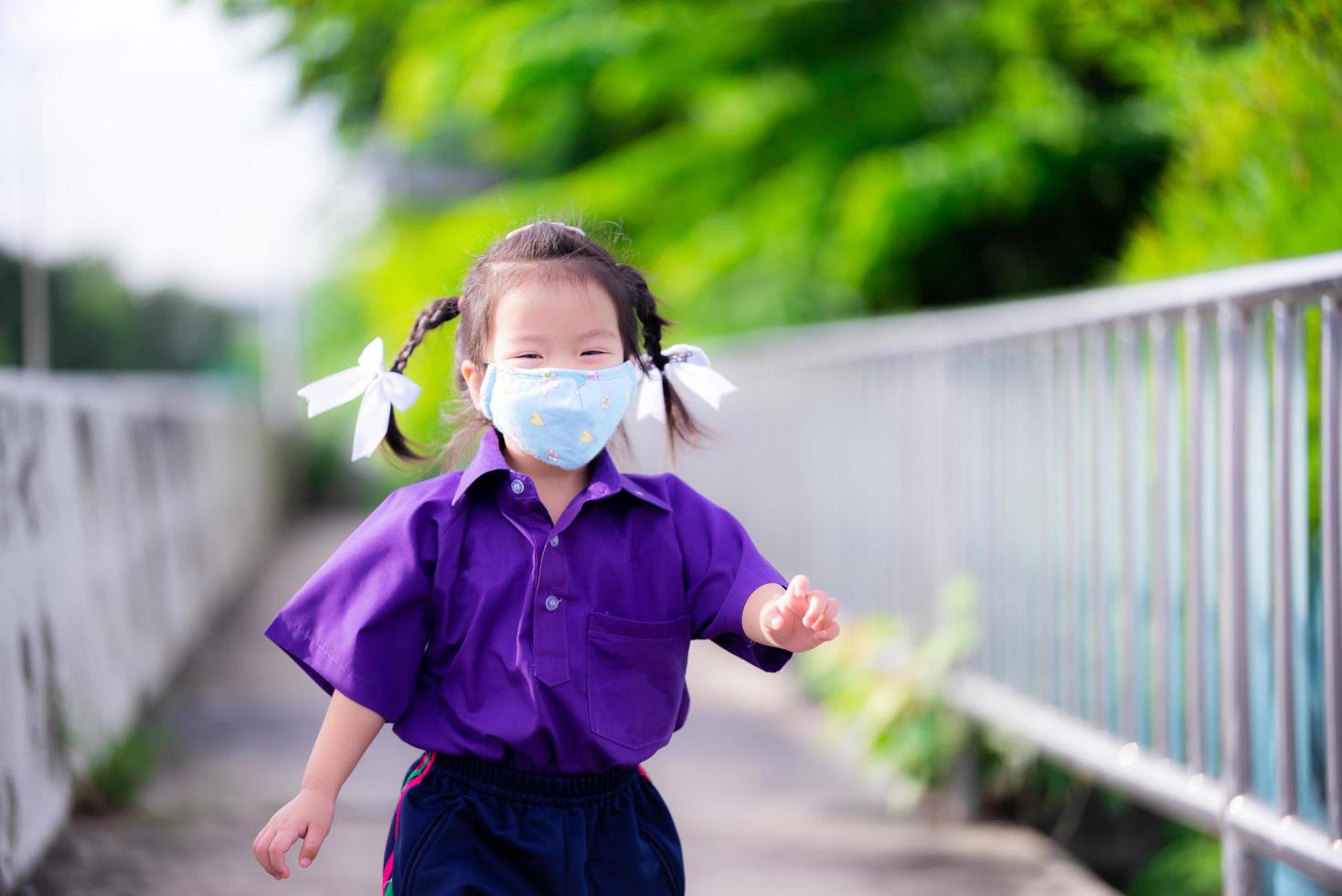Student mit blauer Stoffmaske läuft auf der Straße. Kinder tragen lila Sportuniformen der Schule. Mädchen lächeln süß unter der Maske. Ausbreitung von Coronavirus und giftigem Staub verhindern pm 2.5. Kind 3 Jahre alt foto
