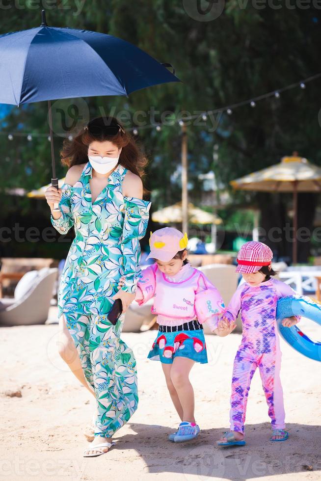 vertikales Bild. schönes Porträt der Familie reisen den Strand. Mutter und Tochter gehen auf Sand. Kind hält Gummiring. Mama breitete einen Regenschirm aus, um sich vor der heißen Sonne zu schützen. Sommerzeit. foto