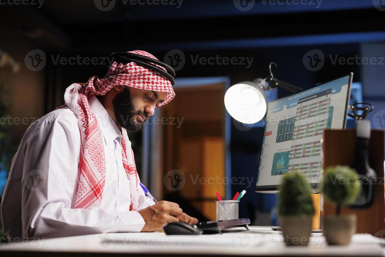 Seitenansicht von ein Muslim Unternehmer mit ein Stift zu schreiben Nieder Anmerkungen empfangen von seine pc Monitor. Bild zeigt an ein gewidmet arabisch Mann mit ein Notizbuch und Desktop zum seine Forschung. foto