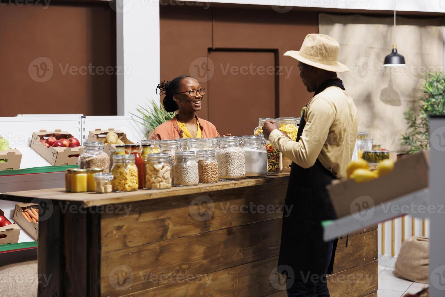 männlich Geschäft Inhaber diskutieren frisch Früchte und Gemüse beim lokal Markt mit ein lächelnd schwarz Frau. Verkäufer bietet an organisch Speisekammer Heftklammern und kunststofffrei Bulk Produkte, fördern gesund produzieren. foto
