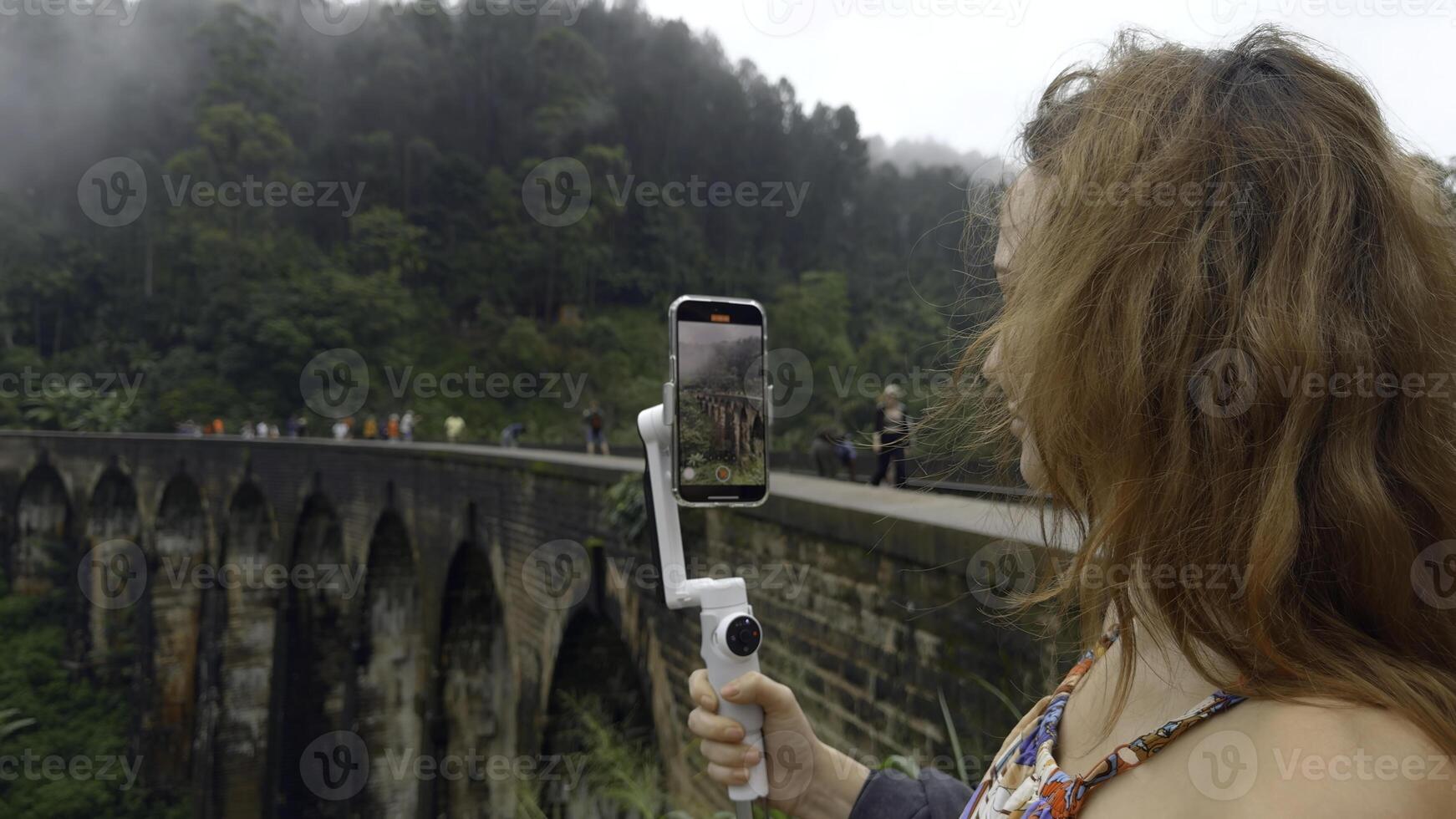 Frau nimmt auf Telefon mit Stein Brücke und Grün Berge. Aktion. schön Frau schießt Blog im tropisch Berge. Frau nimmt schön Ansichten von Urwald und Stein Brücke auf Telefon foto