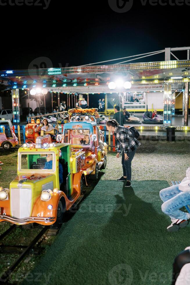 Mama steht in der Nähe von das Kinder Eisenbahn Wagen im das Amüsement Park und sieht aus beim das wenig Mädchen foto