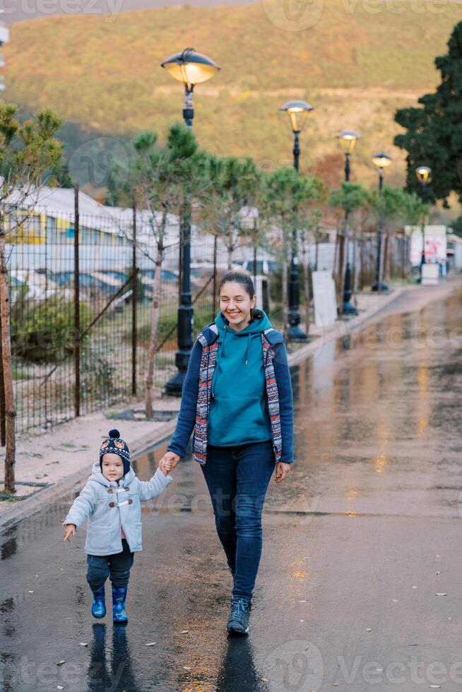 lächelnd Mutter mit ein wenig Mädchen Gehen halten Hände entlang ein nass Straße mit glühend Laternen foto