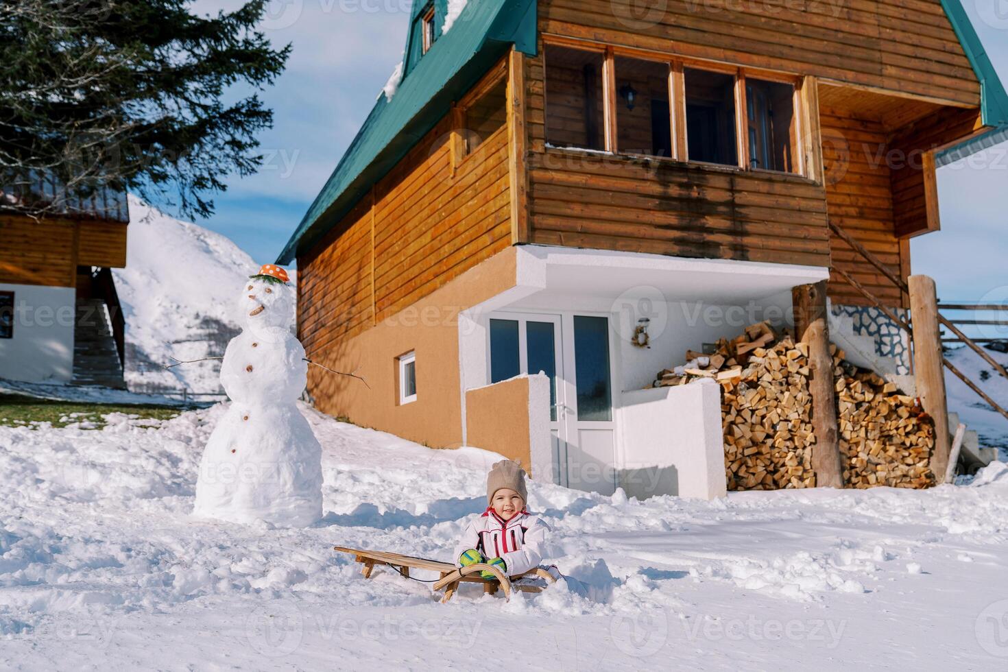 wenig Mädchen sitzt auf ein Schlitten in der Nähe von ein Schneemann Nächster zu ein hölzern Hütte foto
