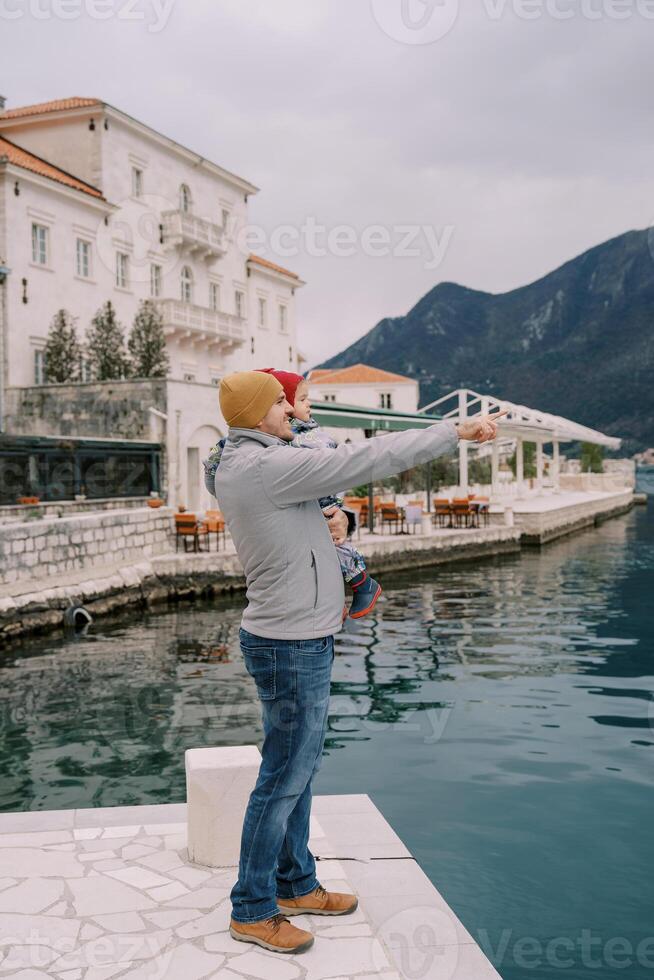 Papa mit ein wenig Mädchen im seine Waffen steht auf das Seebrücke von das alt Stadt, Dorf und Punkte zu das Berge. Seite Aussicht foto