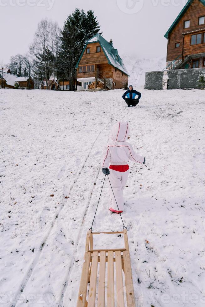 Papa Kniebeugen auf ein Hügel und Uhren ein wenig Mädchen gehen oben mit ein Schlitten auf ein Schnur. zurück Aussicht foto