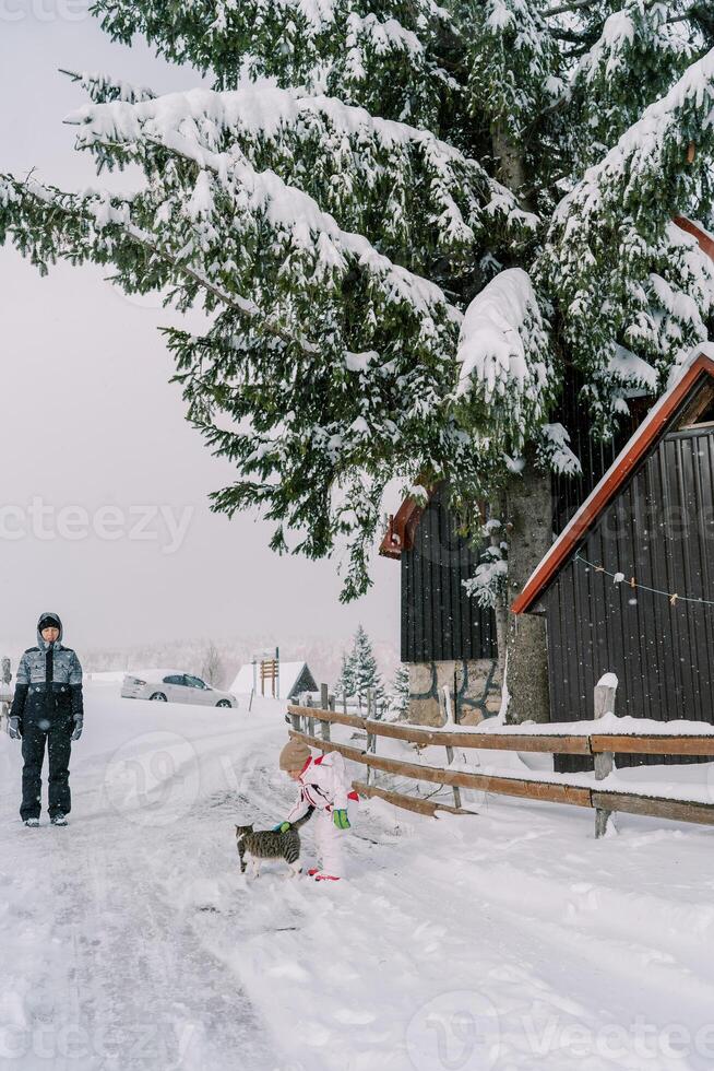 Mama sieht aus beim ein wenig Mädchen streicheln ein Katze während Stehen auf ein schneebedeckt Straße in der Nähe von das Zaun von das Haus foto