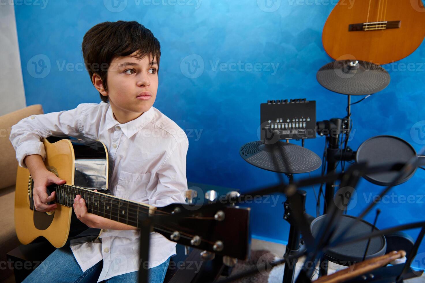 authentisch Porträt von ein Jugendlicher Junge spielen Gitarre im das modern Musik- Studio. elektrisch Gitarre hängend auf ein Blau Mauer in der Nähe von Schlagzeug. Personen. Musik- Lektion. Kinder Bildung und Unterhaltung foto