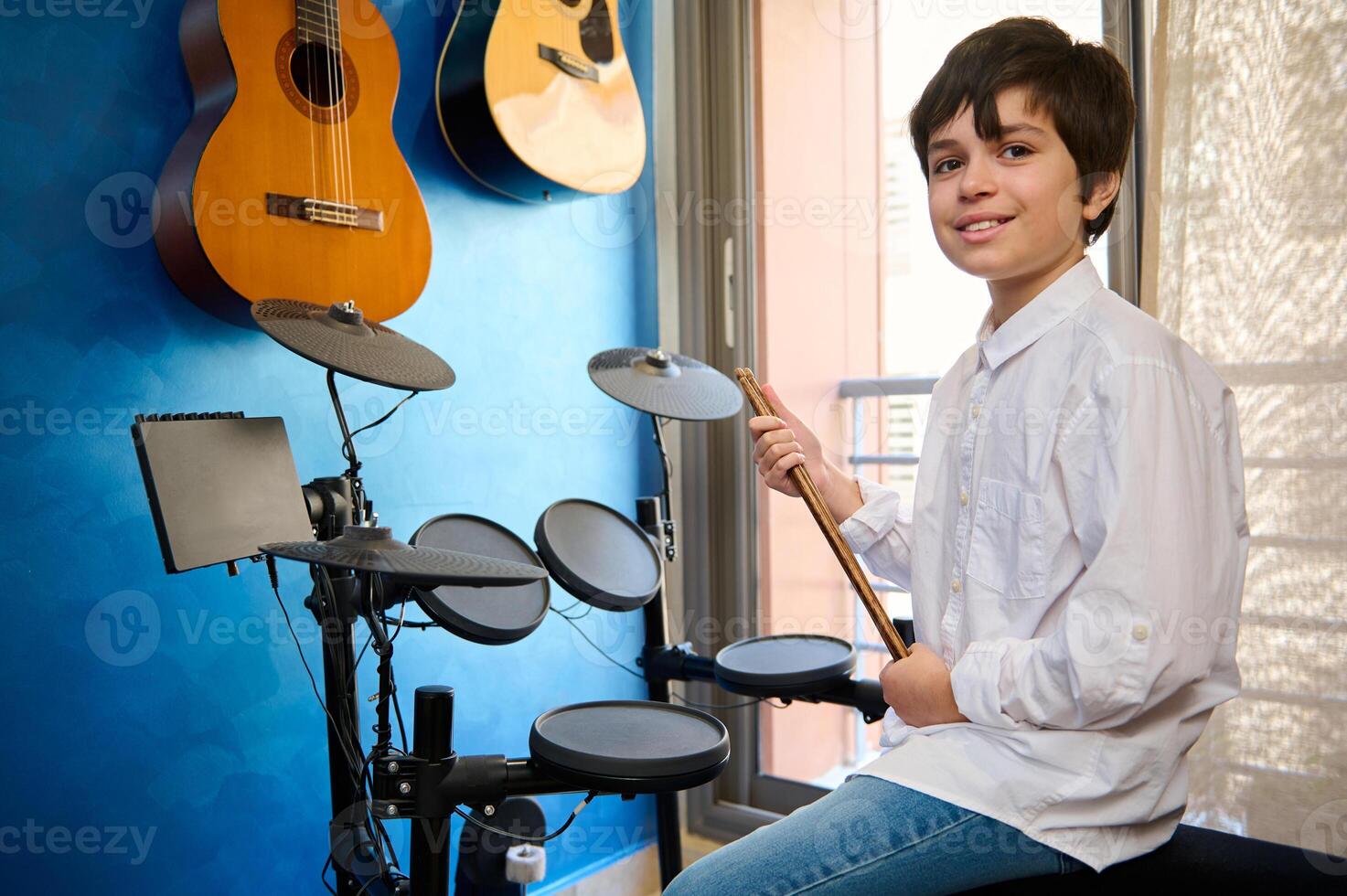 Teenager Junge Schlagzeuger Musiker im Weiß Hemd und Blau Jeans, Sitzung beim Trommel einstellen im seine retro Musik- Studio , halten Trommelstock und lächelnd suchen selbstbewusst beim Kamera. Gitarren hängend auf ein Blau Mauer foto