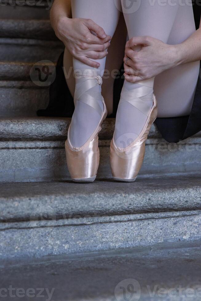 Nahansicht von Ballerina Punkte auf das grau Stein Schritte Hintergrund. tanzen Sitzung auf das Treppe. Ballerina ist Verpackung ihr Hände um ihr Knöchel. Tänzer Füße mit Rosa Ballett Hausschuhe An. Kopieren Raum foto