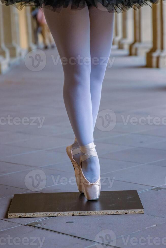 Nahansicht von ein Tänzer Beine balancieren auf das pointe Schuhe. Ballerina tragen Ballett Hausschuhe während Stehen auf ihr Tipps auf das Kopfsteinpflaster Straße. grau retro Türen auf das Hintergrund. selektiv Fokus foto