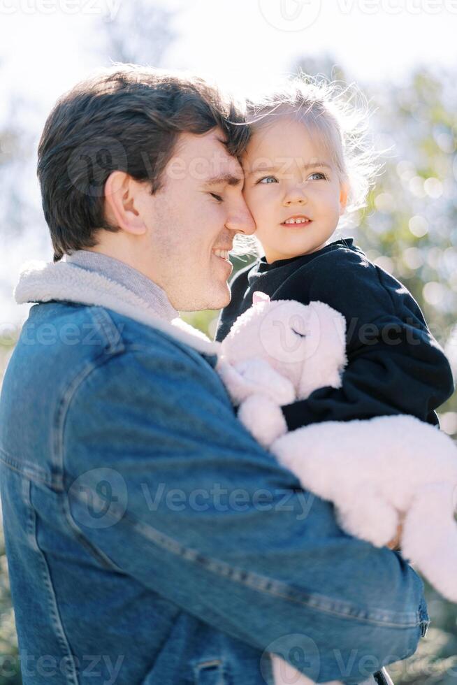 lächelnd Papa berührt seine Nase zu das Wange von ein wenig Mädchen Sitzung im seine Waffen im das Garten foto