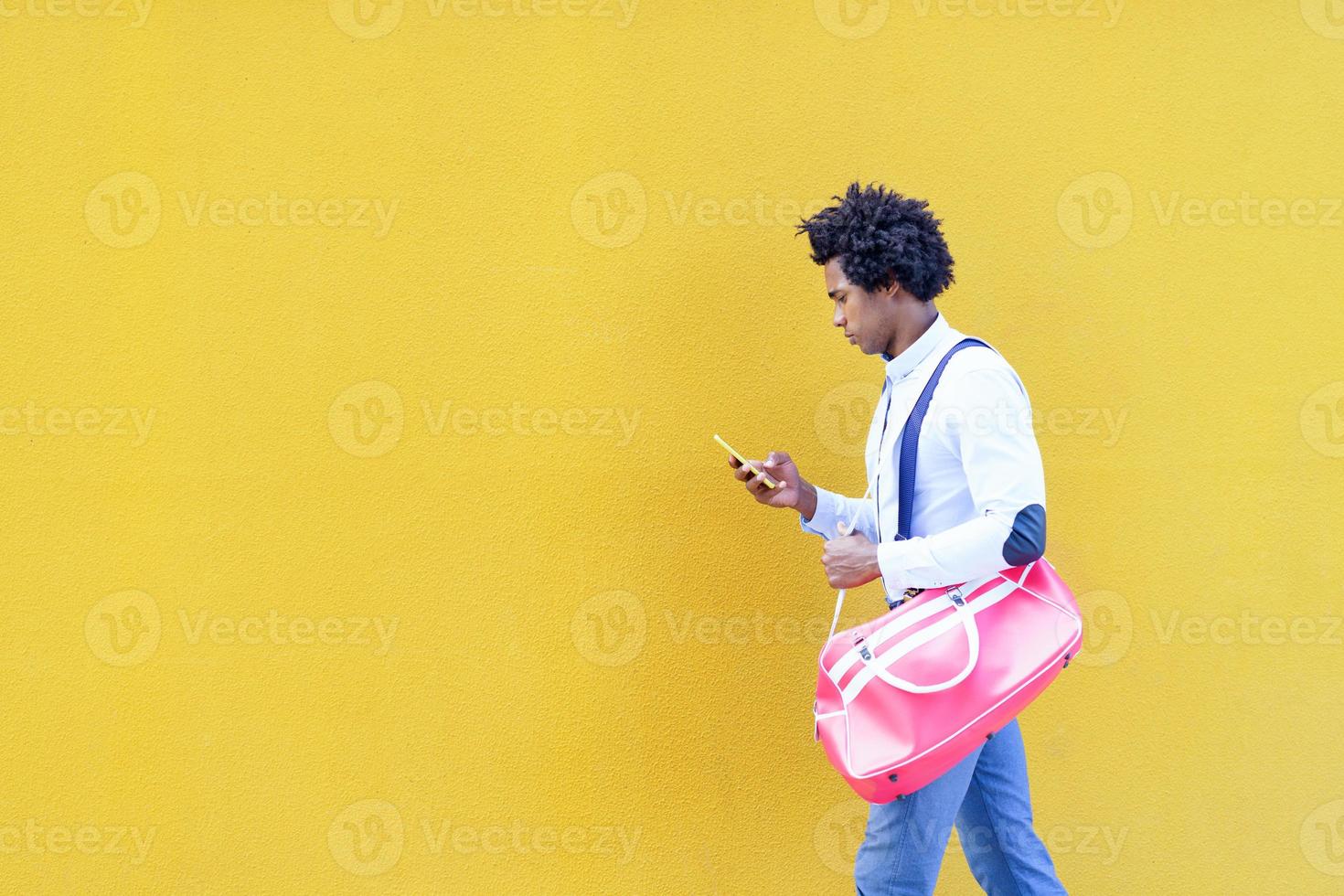 schwarzer Mann mit Afro-Frisur, der eine Sporttasche und ein Smartphone in gelbem Hintergrund trägt. foto