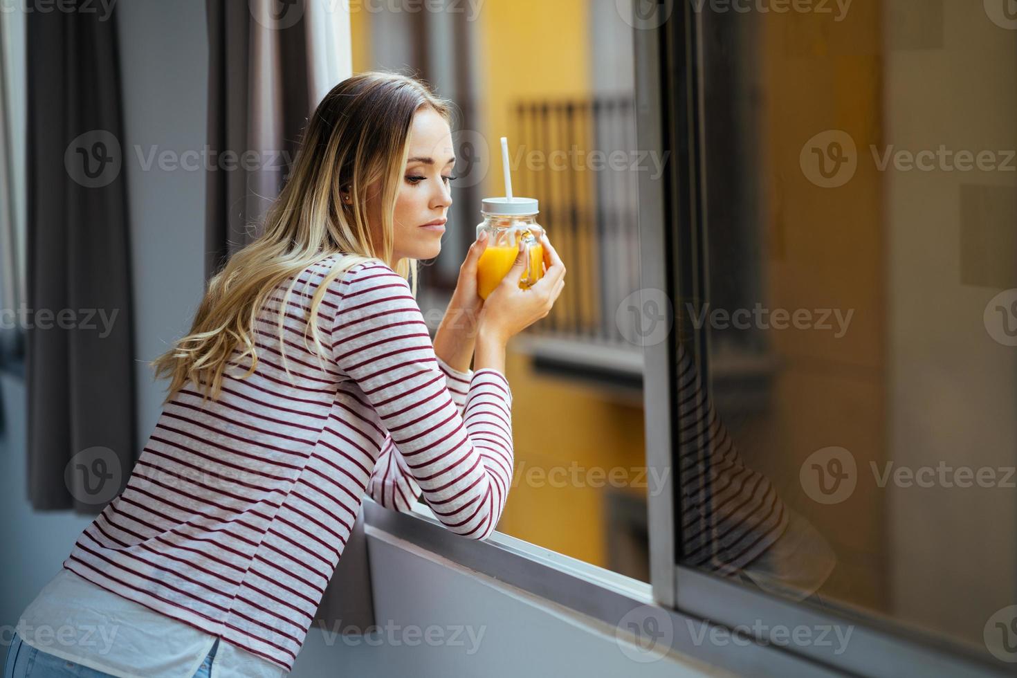 junge Frau trinkt ein Glas natürlichen Orangensaft und lehnt sich aus dem Fenster ihres Hauses. foto