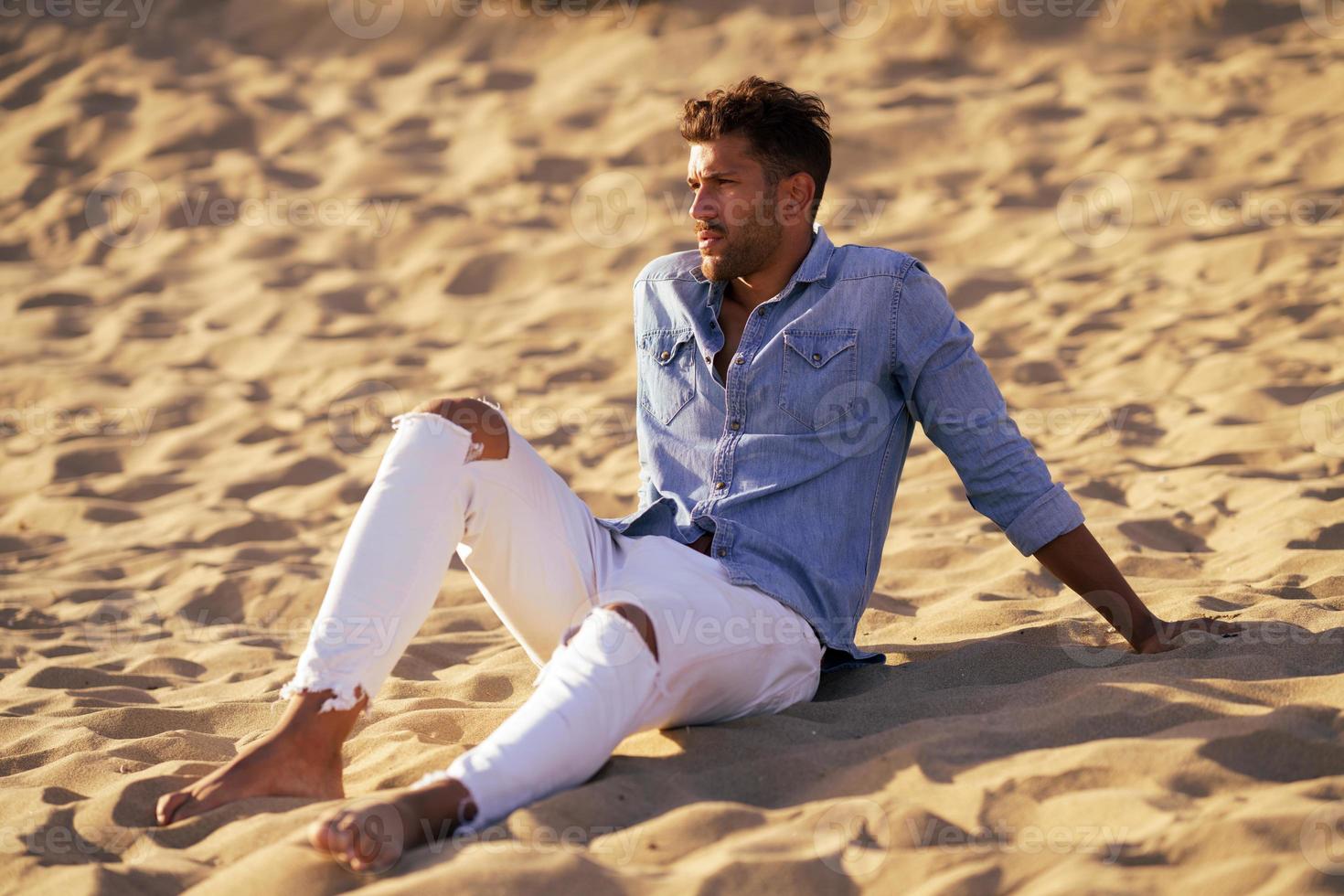 attraktiver Mann, der auf dem Sand des Strandes sitzt foto