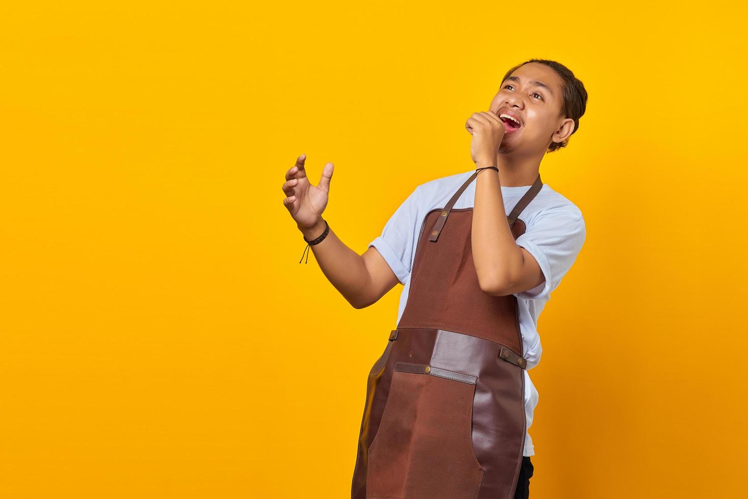 Porträt von fröhlichen asiatischen Jungen, die eine Schürze tragen, wird gesehen, wie sie auf gelbem Hintergrund singen foto