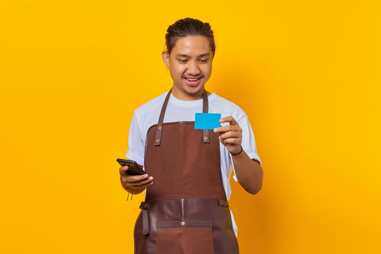 Porträt eines fröhlichen, gutaussehenden Barista, der auf die Kreditkarte schaut und das Smartphone isoliert auf gelbem Hintergrund hält foto
