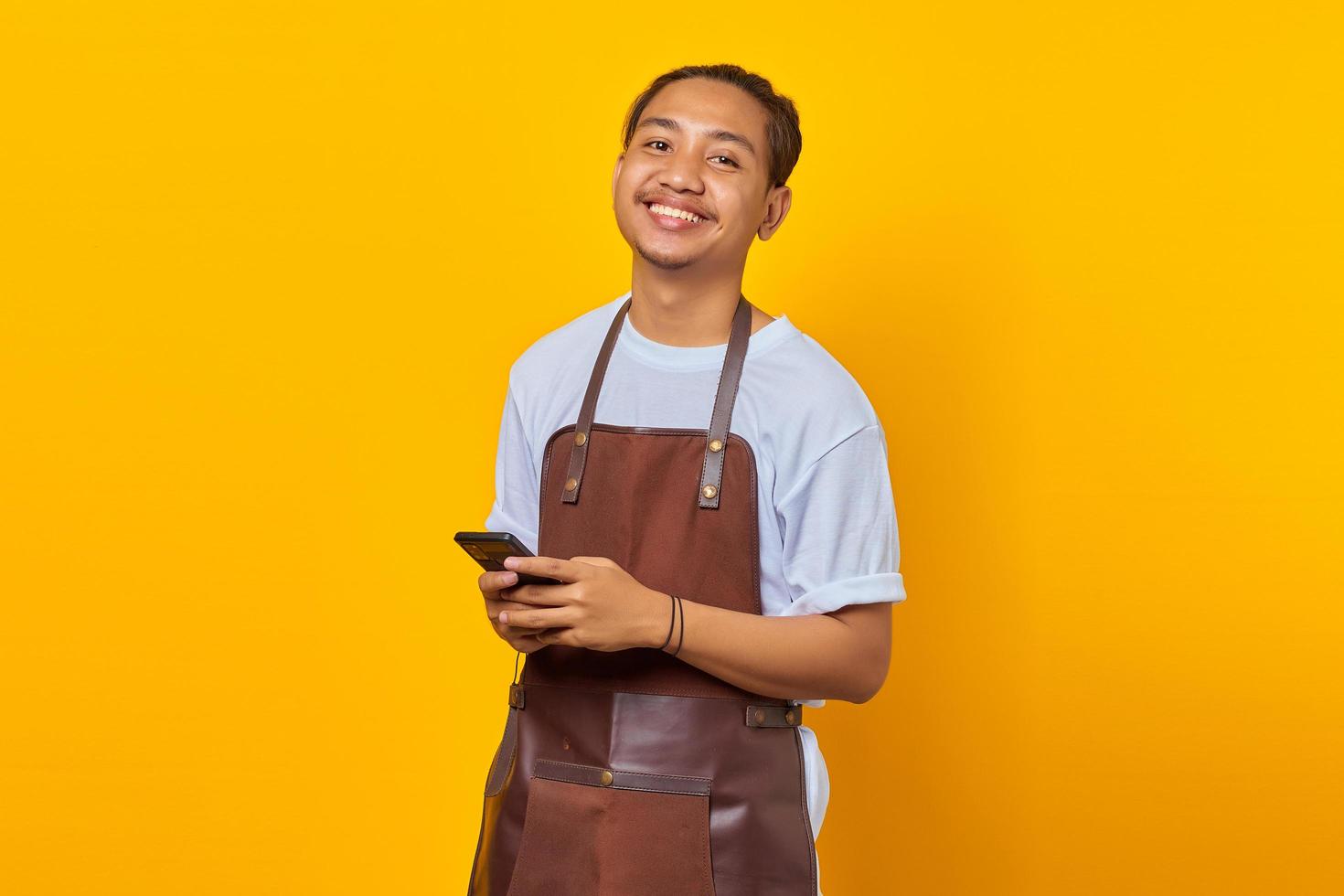 Porträt eines fröhlichen, gutaussehenden Barista, der nach vorne schaut und Smartphone auf gelbem Hintergrund hält foto