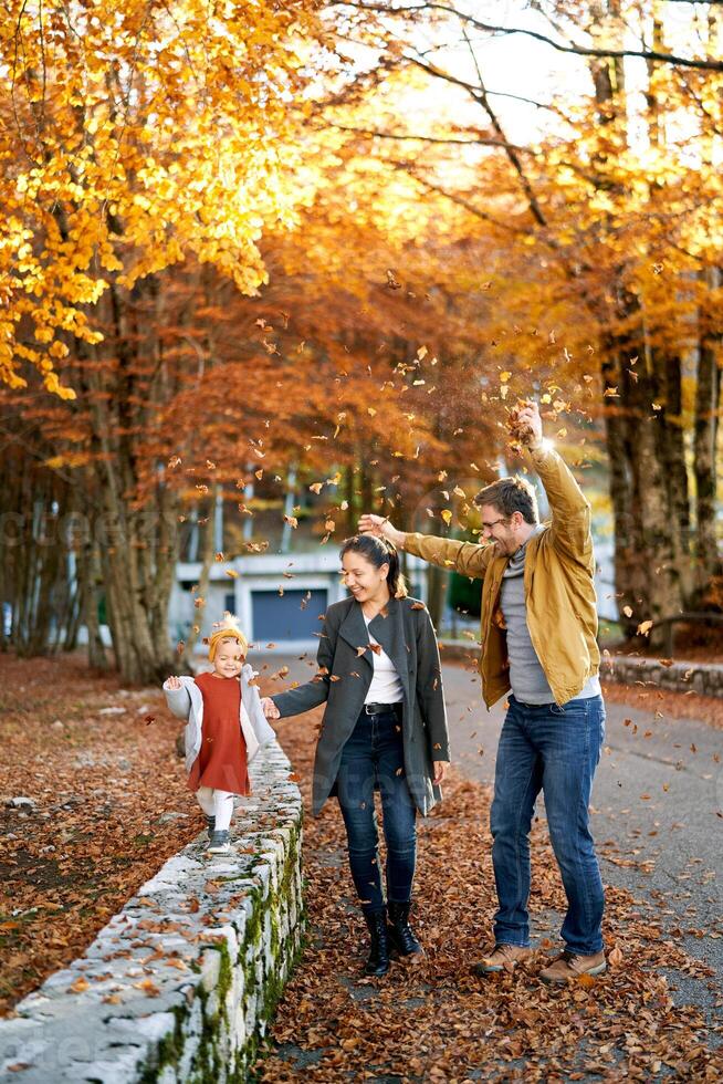 Mama führt ein wenig Mädchen durch das Hand entlang das Zaun im das Park Nächster zu Papa Prise Blätter auf Sie foto