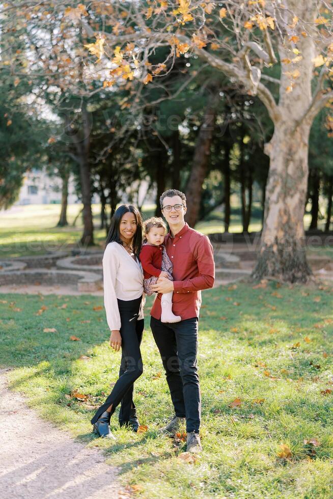 Mama steht Nächster zu Papa halten ein wenig Mädchen im das Park foto