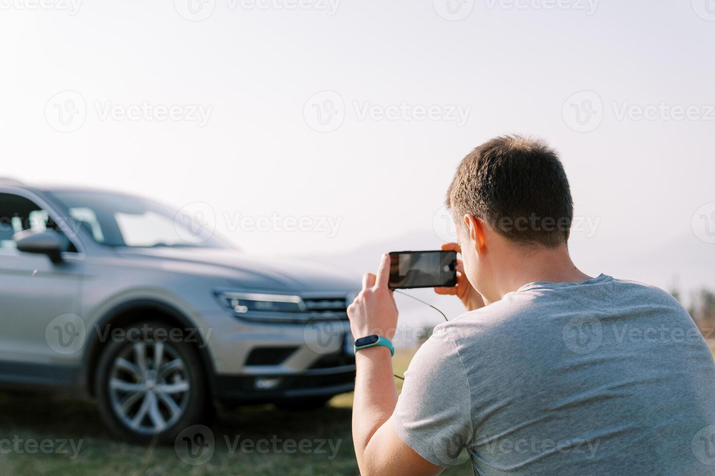 Treiber nimmt ein Bild von seine Auto auf ein Grün Rasen mit ein Smartphone. zurück Aussicht foto