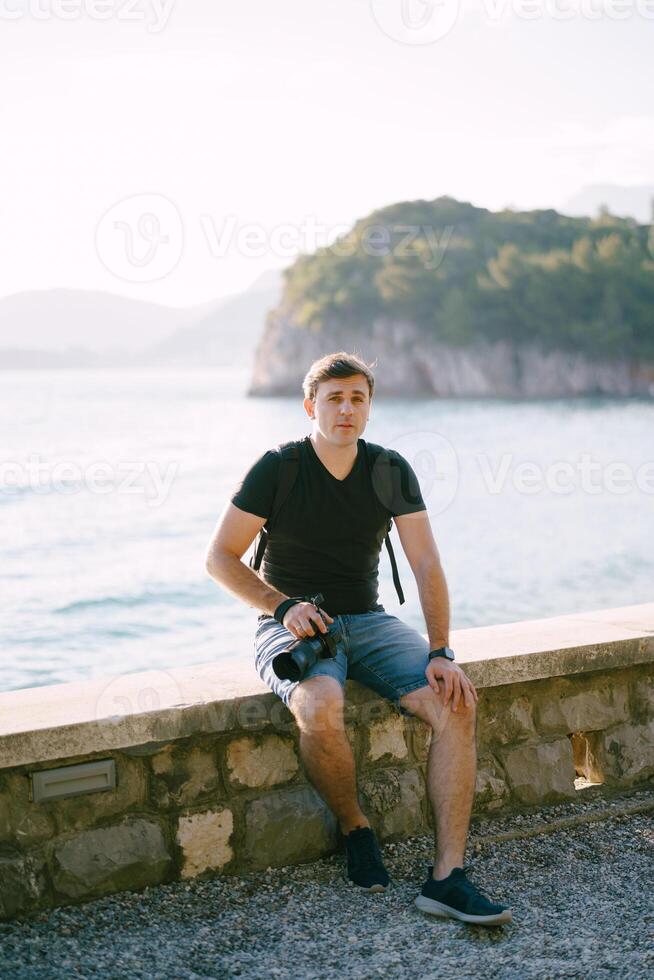 Fotograf mit ein Rucksack sitzt auf ein Stein Zaun durch das Meer foto