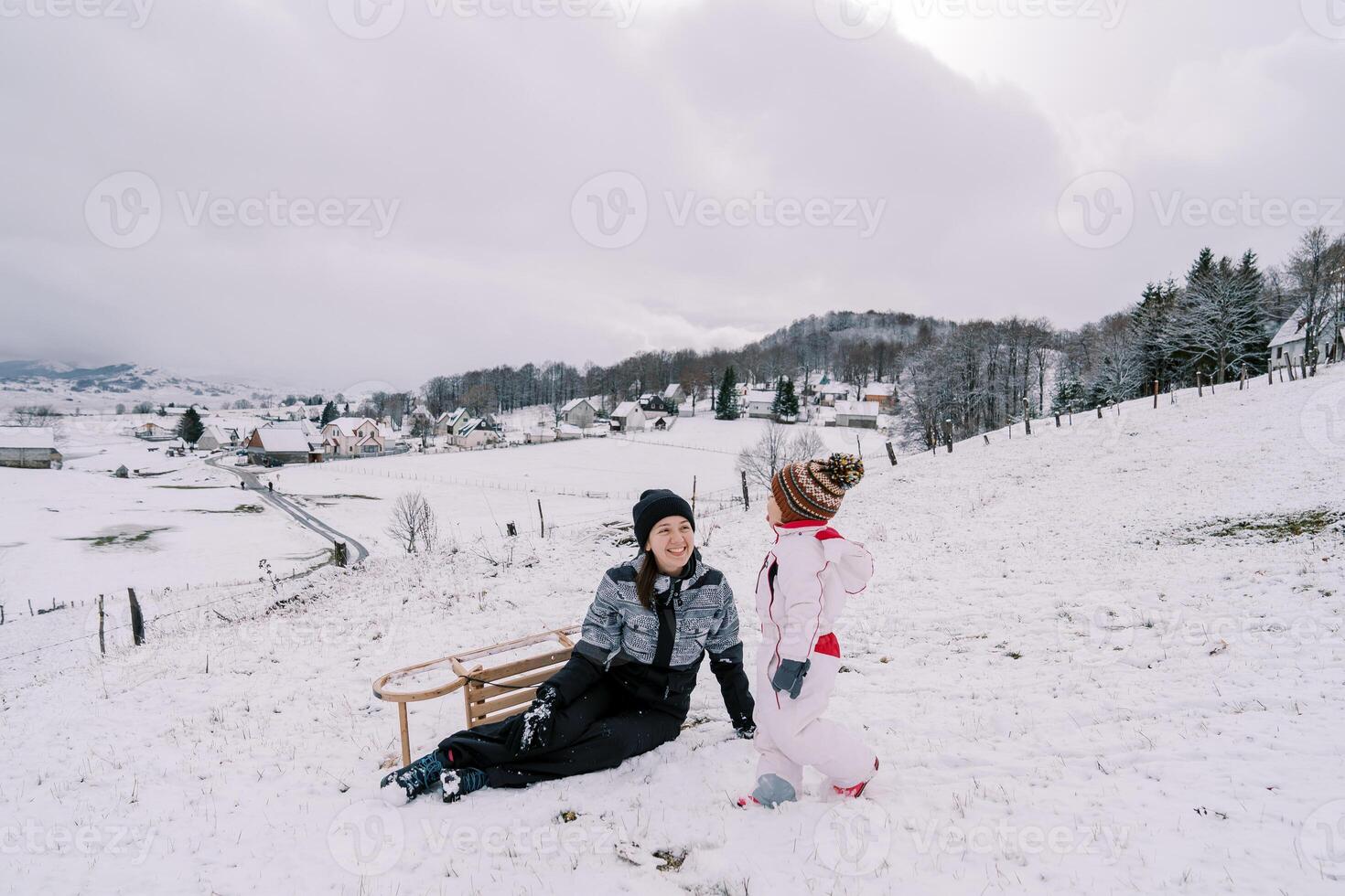 wenig Mädchen Spaziergänge entlang ein schneebedeckt Hügel gegenüber ihr Sitzung lächelnd Mutter mit ein Schlitten gedreht auf es ist Seite foto