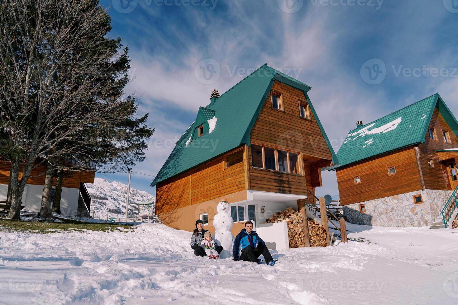 Papa sitzt Nächster zu Mama mit ein wenig Mädchen auf ihr Runde in der Nähe von ein Schneemann im das Schnee Nächster zu ein hölzern Hütte foto