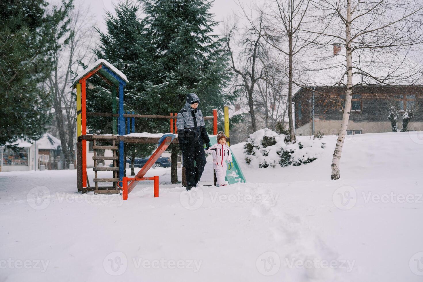 wenig Mädchen und ihr Mutter gehen halten Hände von ein bunt schneebedeckt rutschen durch das Park unter Schneefall foto