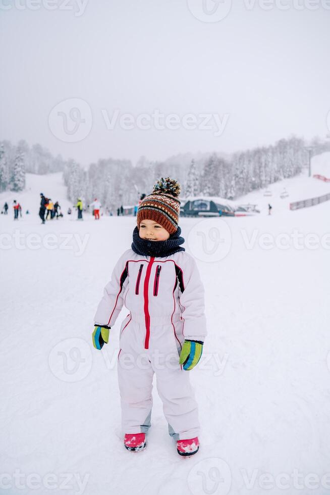 wenig lächelnd Mädchen steht auf ein schneebedeckt Ski Steigung gegen das Hintergrund von Skifahrer foto