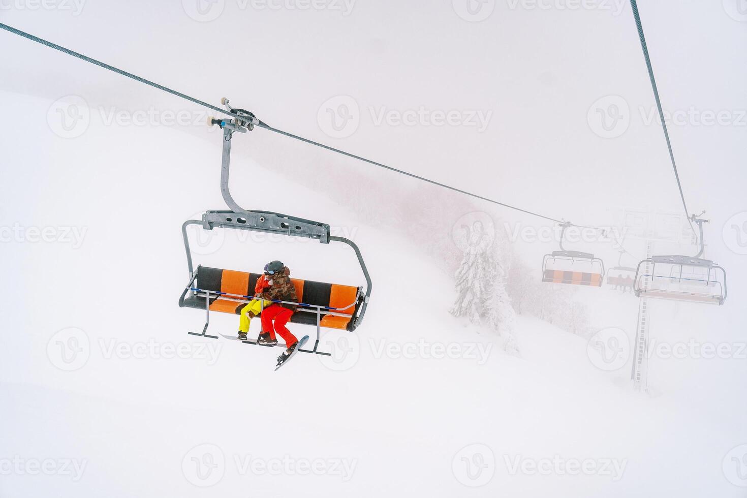 Paar im bunt Ski Anzüge Fahrten umarmen auf ein Sessellift durch das Nebel zu das Berghang foto