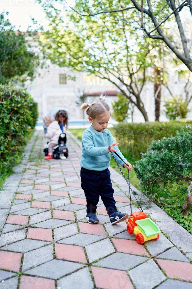 wenig Mädchen mit ein bunt Spielzeug Auto auf ein Zeichenfolge steht auf ein gepflastert Pfad im das Garten foto