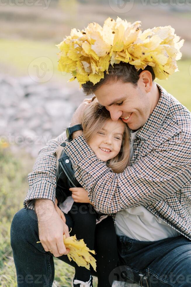 lächelnd Papa im ein Kranz von Gelb Blätter Umarmungen ein wenig Mädchen Sitzung auf seine Runde foto