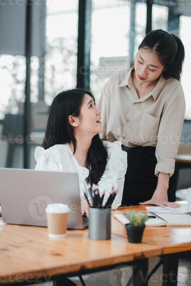 zwei Fachmann Frauen engagieren im ein Kollaborativ Diskussion beim ein gut organisiert Büro Schreibtisch, mit einer Sitzung und das andere Stehen, Teilen ein Moment von freundlich Interaktion. foto