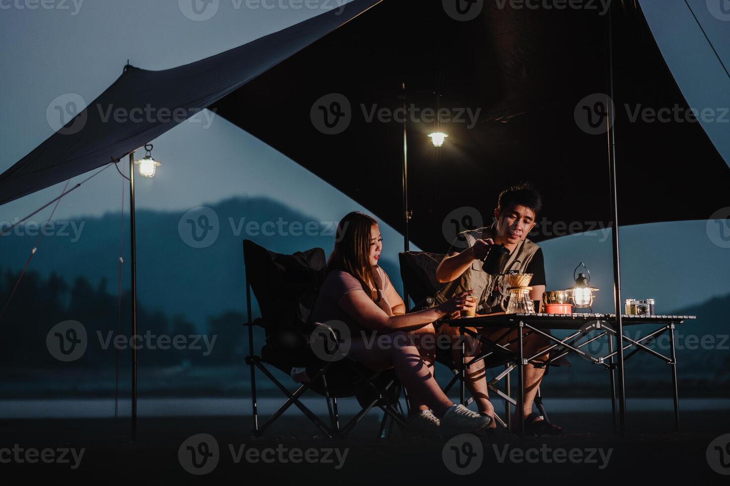 wie Nacht Stürze, ein Paar schmeckt ihr Kaffee und genießt intim Konversation beim ihr gut beleuchtet Seeufer Campingplatz. foto