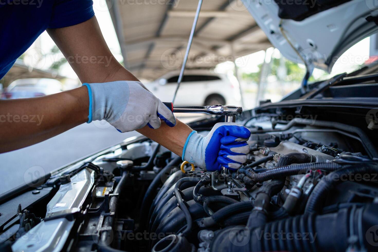 ein Mann ist Arbeiten auf ein Auto Motor. er ist tragen Blau Handschuhe und ein Blau Shirt. das Auto ist geparkt unter ein Dach foto