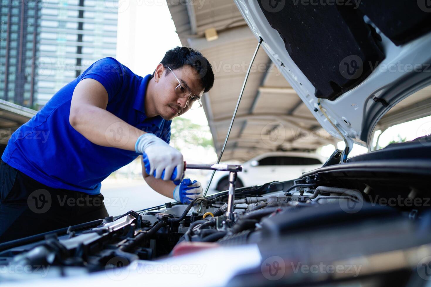 ein Mann ist Arbeiten auf ein Auto Motor. er ist tragen ein Blau Hemd und Handschuhe. das Auto ist geparkt unter ein Gebäude foto