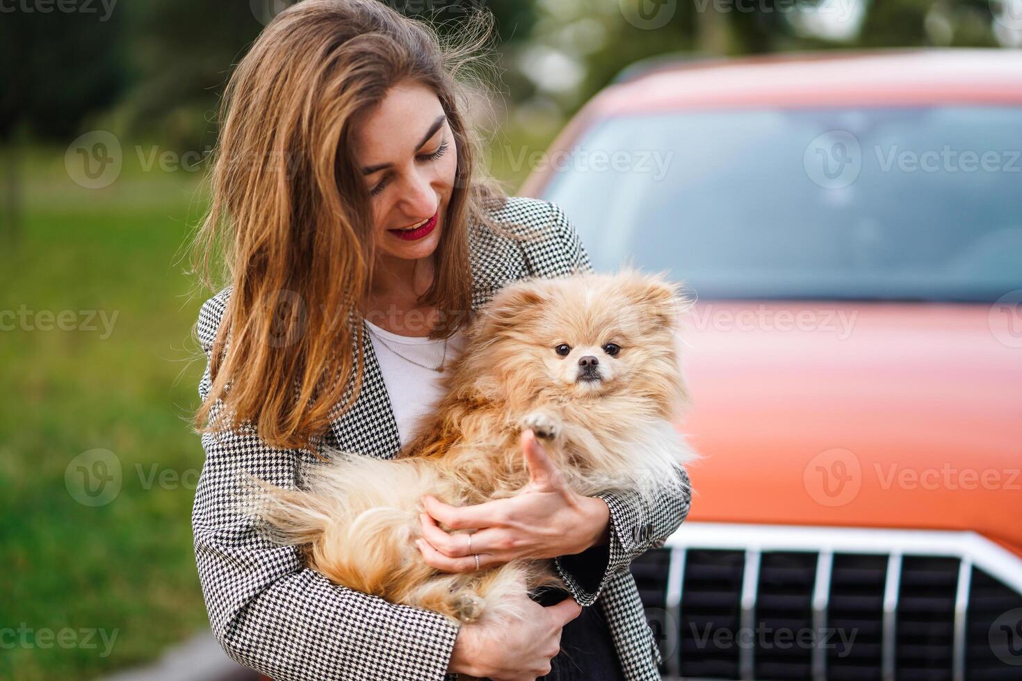 lächelnd Frau halten ein flauschige pommerschen Hund in der Nähe von ein rot Auto im das Park foto