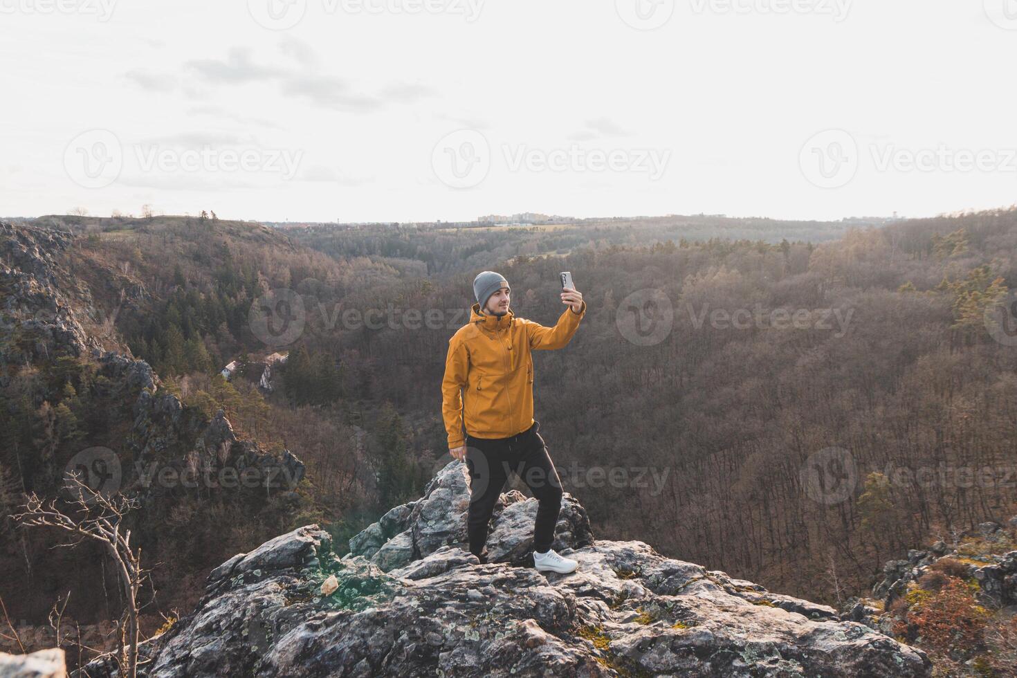 Reisender im ein Gelb Jacke Stehen auf das Kante von ein Cliff nehmen ein Selfie auf seine Handy, Mobiltelefon Telefon beim Sonnenuntergang. Abonnieren sarka Schlucht, Prag foto