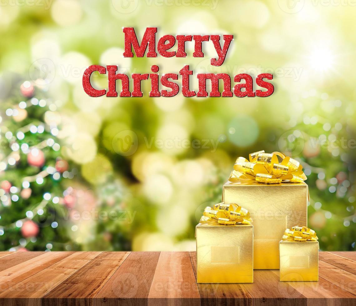 Frohe Weihnachten rotes Glitzerwort und goldenes Geschenk auf braunem Holztisch mit Weihnachtsbaum verschwommenes Bokeh foto