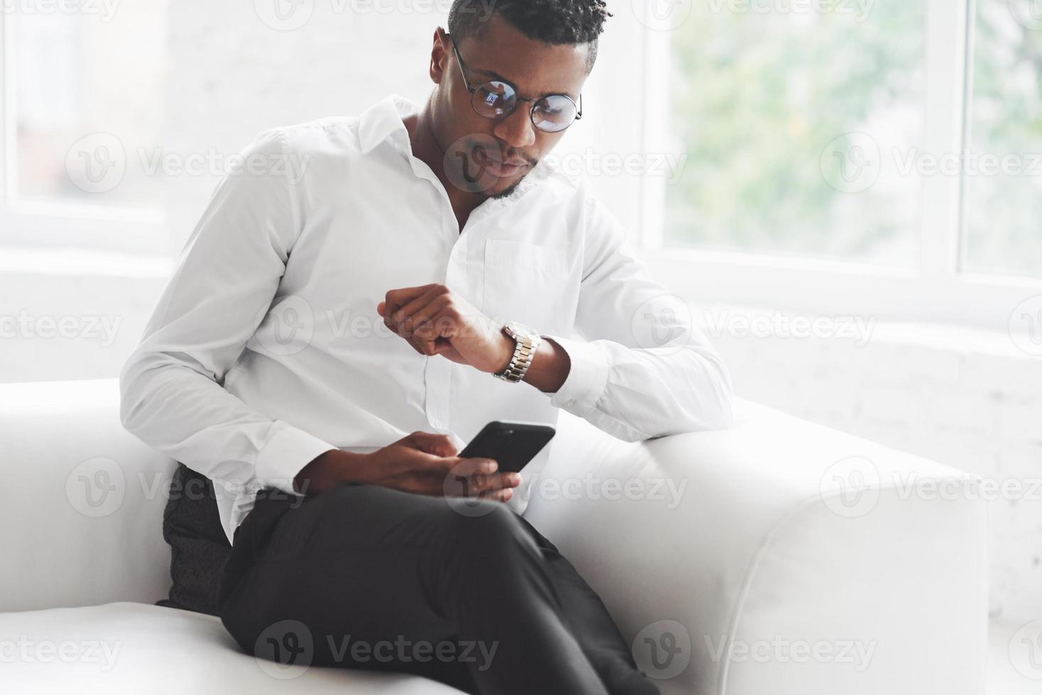 neuer Deal kommt. die Zeit überprüfen. junger Afro-Amerikaner in Bürokleidung sitzt auf dem weißen Sofa foto