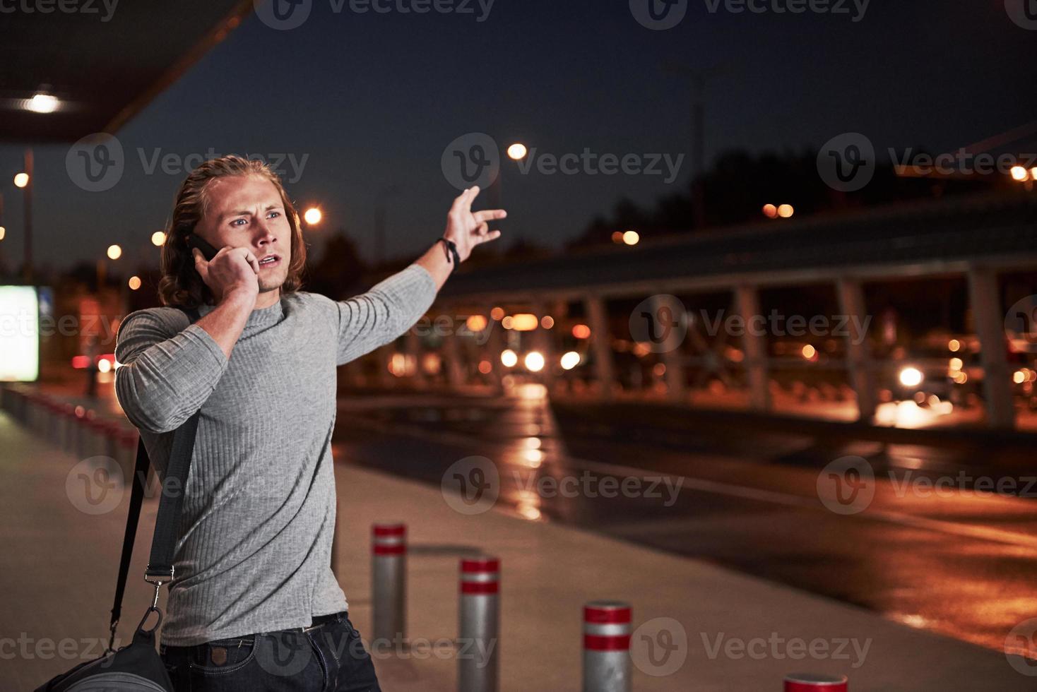 aktive Handbewegungen. junger, stilvoller Mann, der durch die Nachtstraße in der Nähe der Straße geht und ein Gespräch am Telefon führt foto