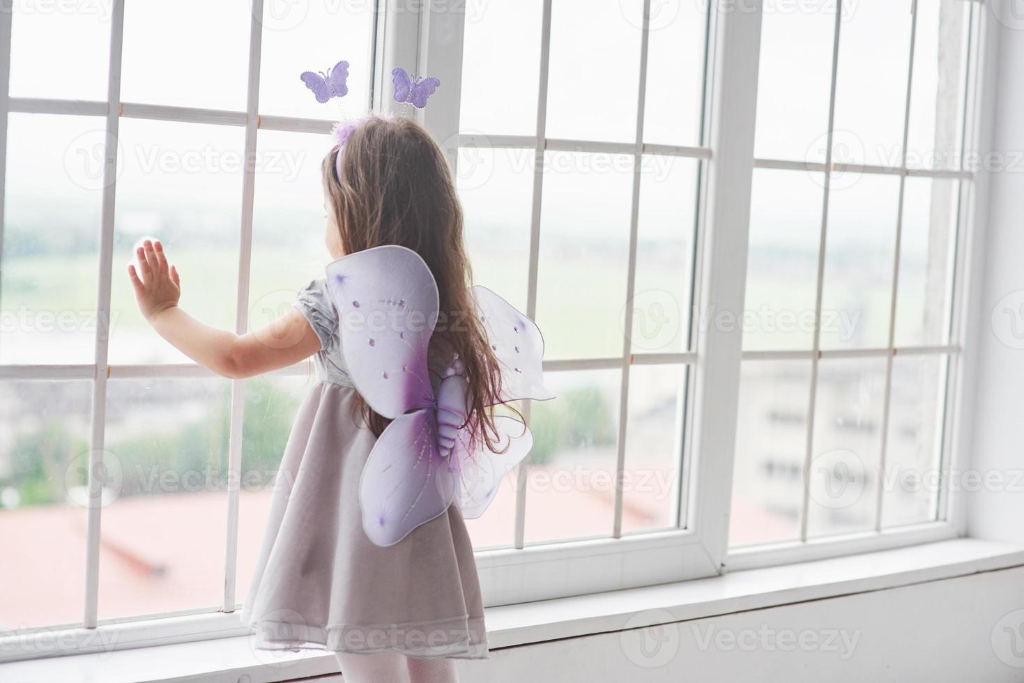 nettes kleines Mädchen in der Märchenkleidung, das neben den Fenstern steht und nach draußen schaut foto