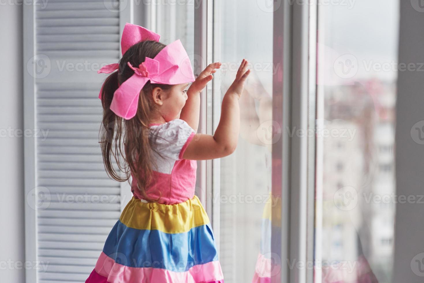 das Glas berühren. Foto eines jungen süßen Mädchens in bunter Kleidung, das in der Nähe des Fensters steht und nach draußen schaut