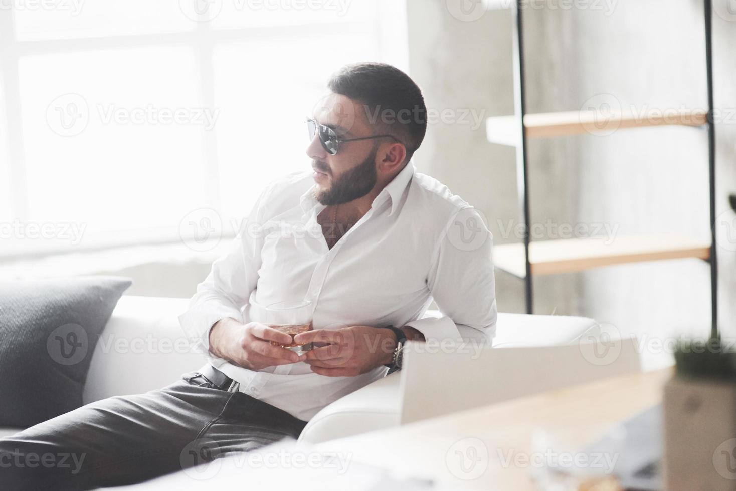 Foto von der Seite. Foto eines jungen bärtigen Geschäftsmannes mit Sonnenbrille und Whisky in der Hand sitzen auf dem weißen Sofa