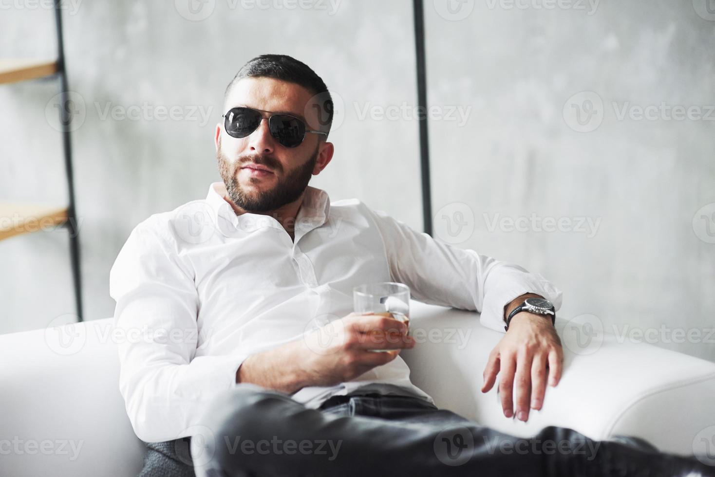 Vorderansicht. Foto eines jungen bärtigen Geschäftsmannes mit Sonnenbrille und Whisky in der Hand sitzen auf dem weißen Sofa