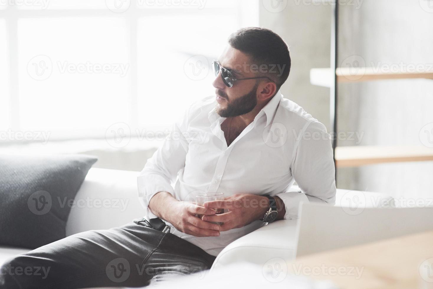 Zeit haben, einige Dinge zu planen. Foto eines jungen bärtigen Geschäftsmannes mit Sonnenbrille und Whisky in der Hand sitzen auf dem weißen Sofa