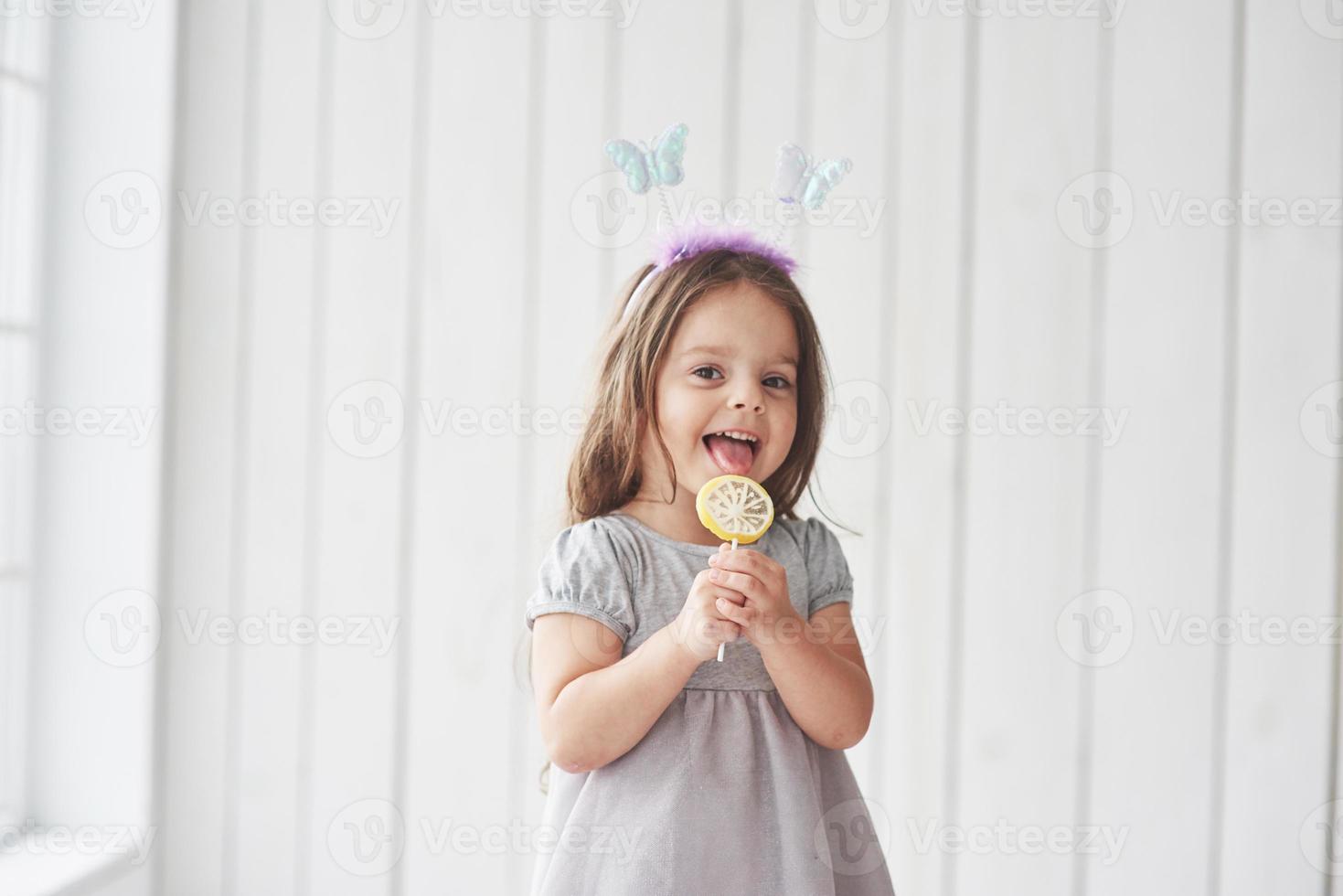 lächeln und Freude empfinden. hübsches kleines Mädchen, das im weißen Raum die gelben Süßigkeiten mit Spielzeugschmetterlingen auf dem Kopf isst foto