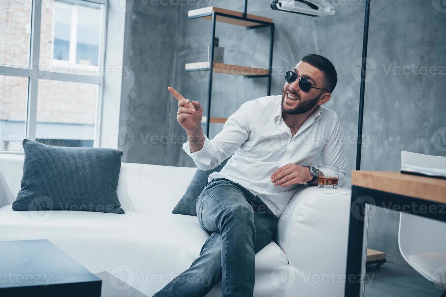 lächelt und zeigt einen Zeigefinger. junger kurzhaariger Mann mit Sonnenbrille sitzt auf der Couch im Büro foto