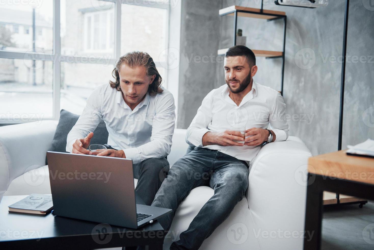 Mann mit langen Haaren zeigt seine Arbeit. Zwei Geschäftsleute, die vor einem Laptop sitzen und über ihre Pläne sprechen foto